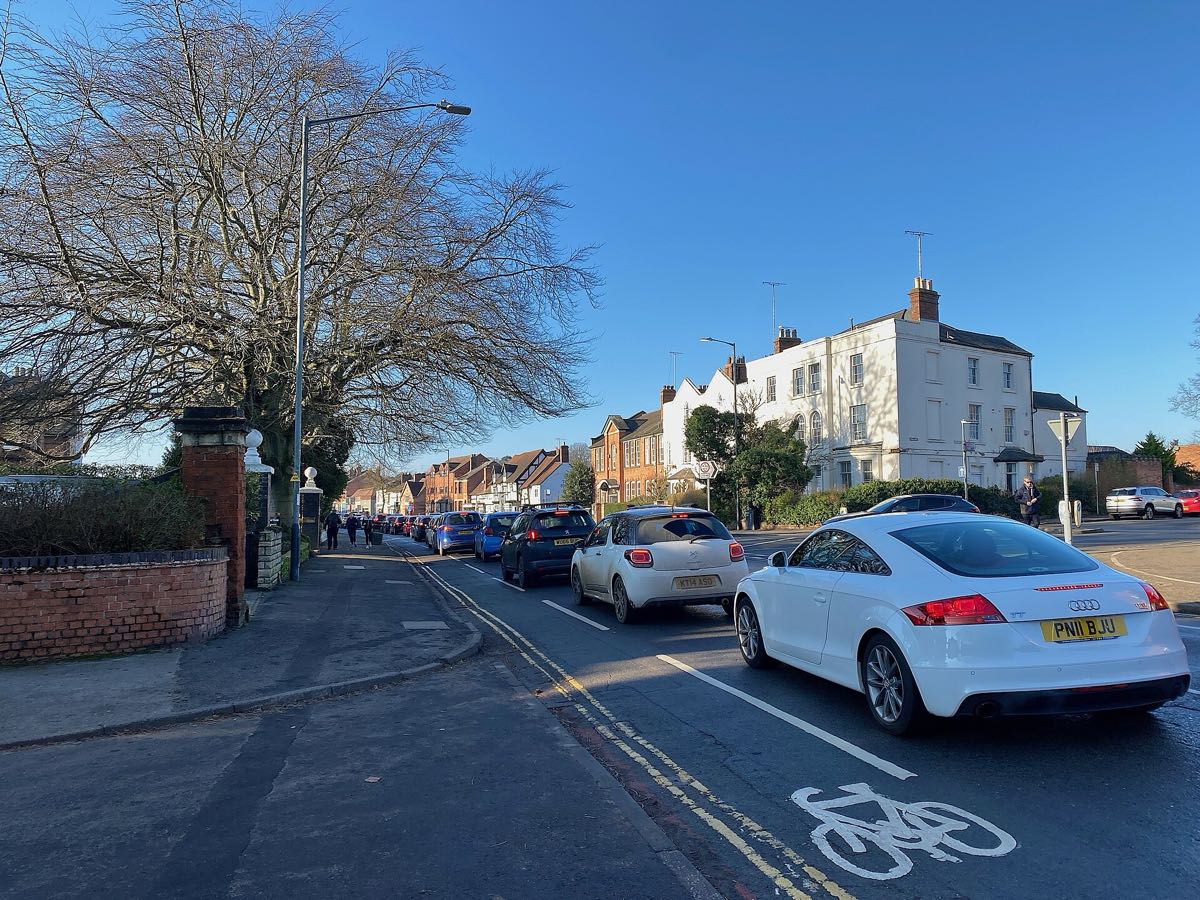A queue of cars on a sunny English street, the nearest showing a yellow rear number plate