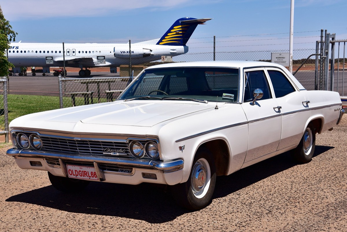 A white 1966 Chevrolet saloon parked at an airfield, its steering wheel visible on the right through the windscreen