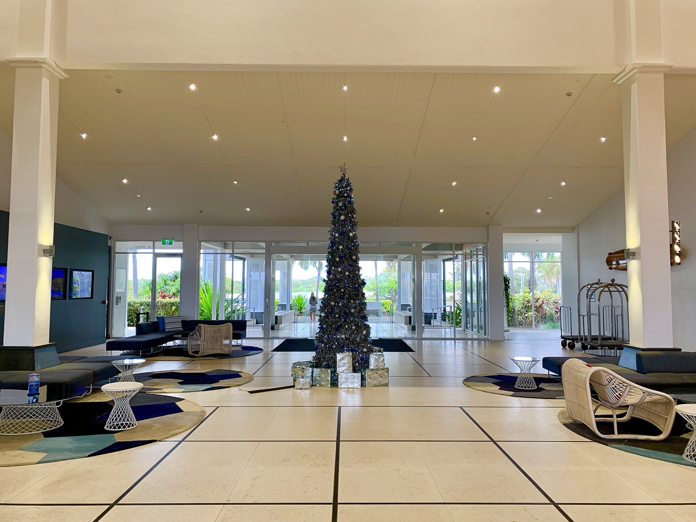 Un gran árbol de Navidad con bolas azules y plateadas en el vestíbulo de un hotel, con sol y palmeras tras los cristales