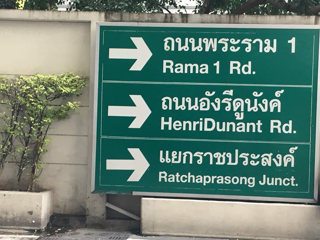 Large green Bangkok street sign with three Thai road names, each with its English transliteration and a white arrow