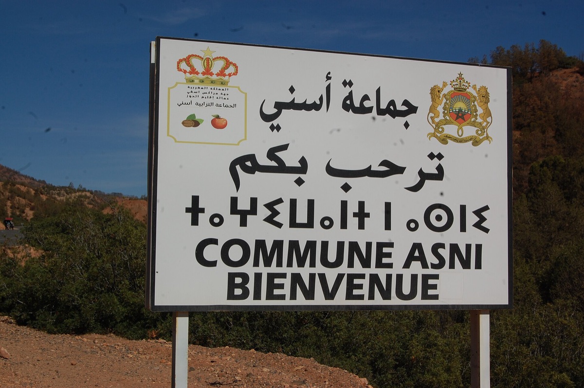 White roadside welcome sign for the commune of Asni, with Arabic, Tifinagh and French stacked on three lines