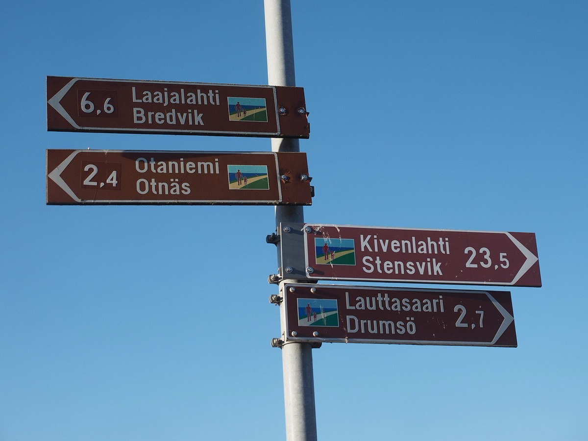Four brown direction signs on one pole, each with a Finnish place name above its Swedish name, against a blue sky