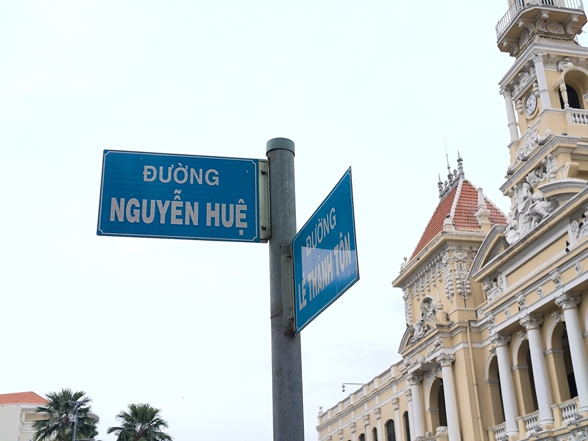 Two blue street signs on one pole reading ĐƯỜNG NGUYỄN HUỆ and ĐƯỜNG LÊ THÁNH TÔN by a yellow building