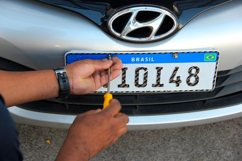A worker screwing a new Mercosur number plate onto a silver car, with BRASIL and the Brazilian flag in the blue top band