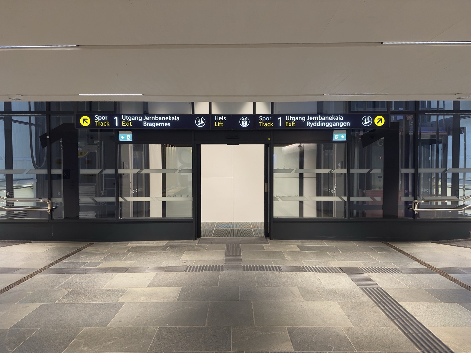 A station sign above glass doors reading Spor 1 Utgang Jernbanekaia Bragernes, with Track and Exit in English underneath